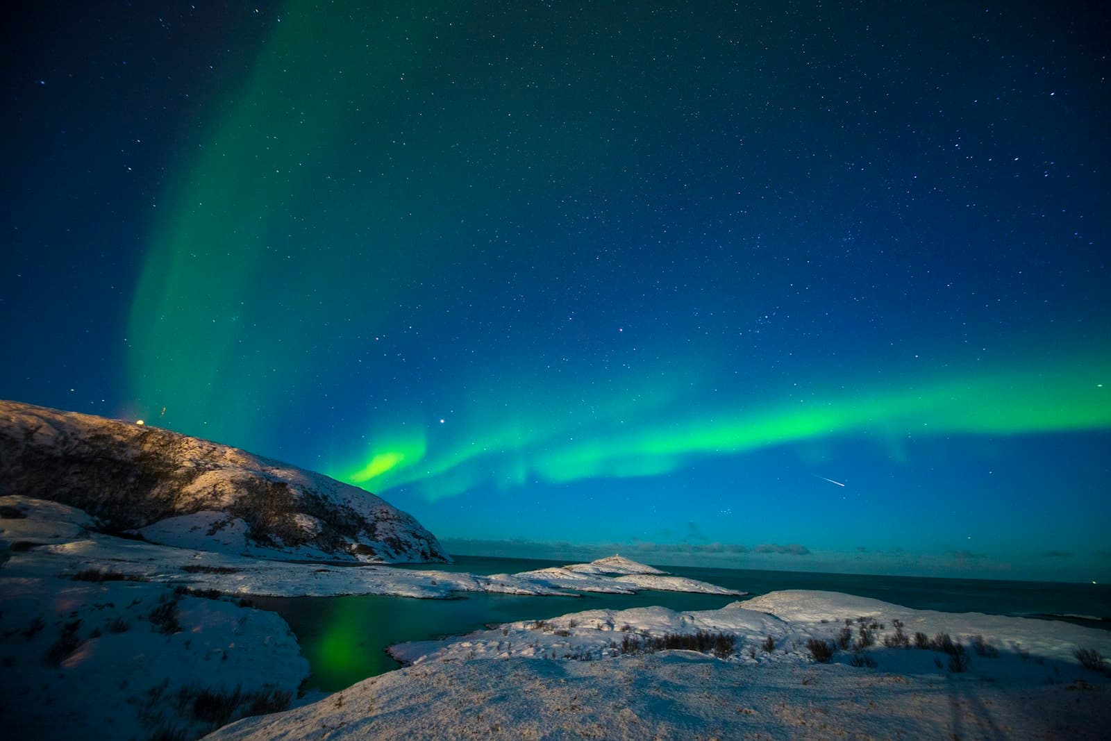 A green aurora band over a snow-covered shoreline and open water in Arctic Norway
