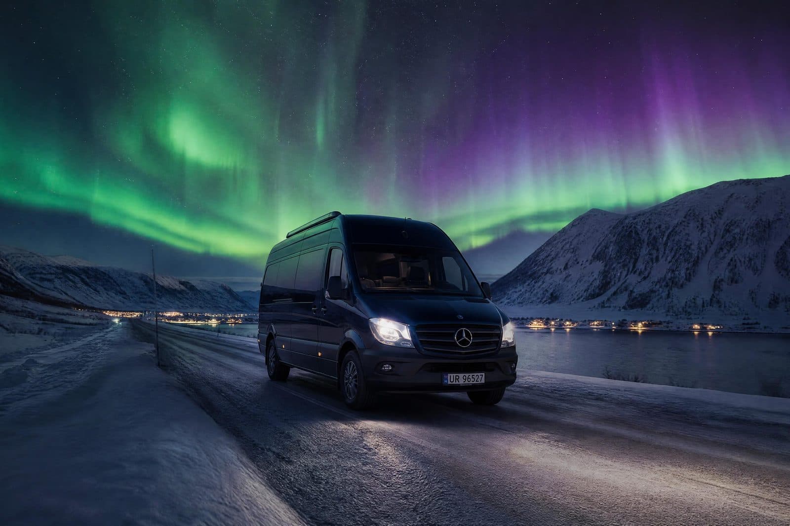 A dark passenger van with its headlights on, on a snowy coastal road under a green aurora