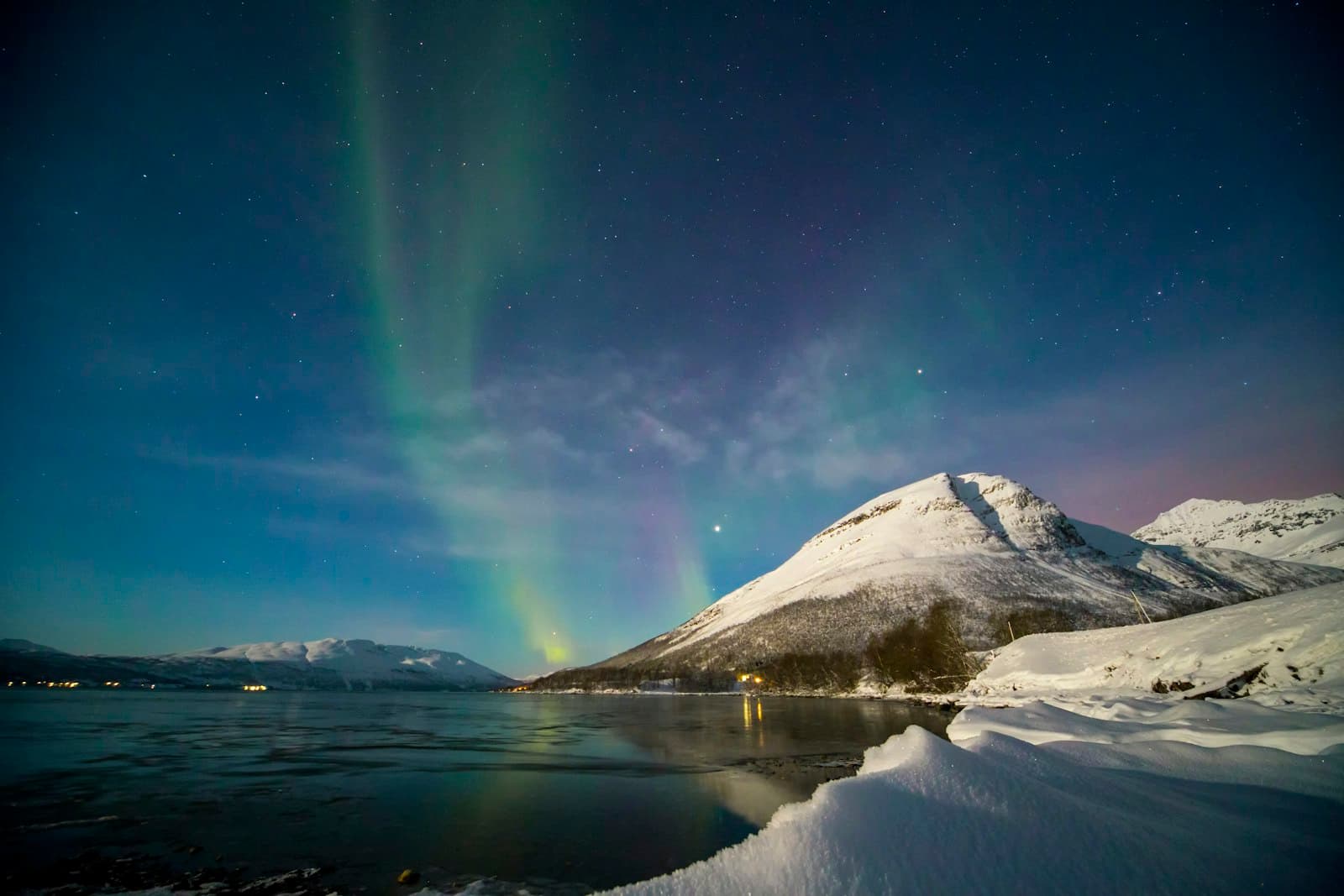An aurora rising behind a snow-covered mountain across a still fjord, with village lights along the shore